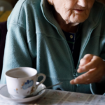 An elderly person in a teal fleece jacket sits at a table with a teacup and saucer, reaching toward the cup.