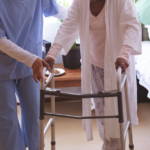 Caregiver assists an elderly person using a walker in a bedroom setting.