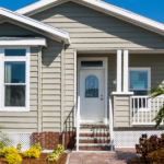 Front view of a beige ranch-style house with white trim, brick foundation, and a small porch; a 'For Sale' sign is in the right window.
