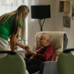 Caregiver in a green shirt hands a cup to an elderly woman in a red sweater sitting in a cushioned chair at home. Option 2: A caregiver leans over to offer a drink to an older woman in a cozy living room. Option 3: A nurse or aide helps an elderly woman relax in a recliner, presenting a cup. Option 4: In a bright living room, a caregiver assists an elderly woman with a beverage. Option 5: An elderly woman receives care from a companion as she sits in a comfortable armchair.